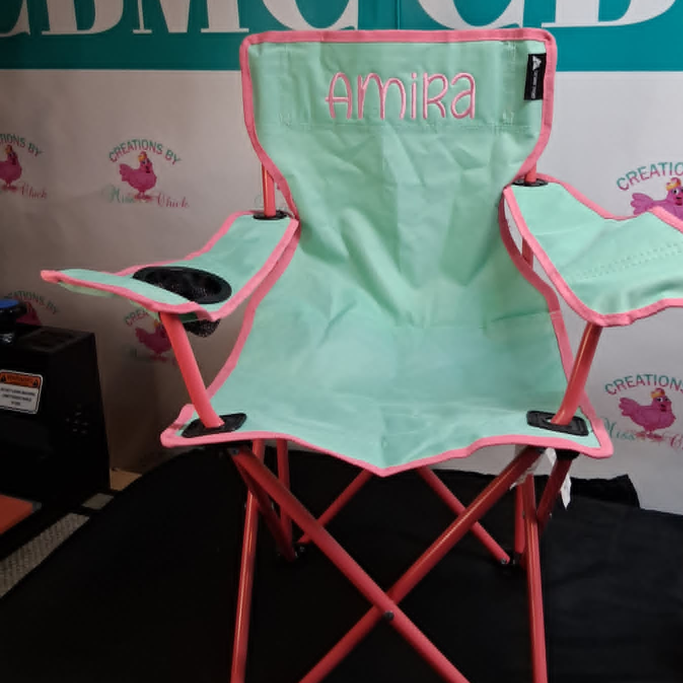 Foldable chair with pink frame and green seat, personalized with 'Amira', against a branded backdrop.